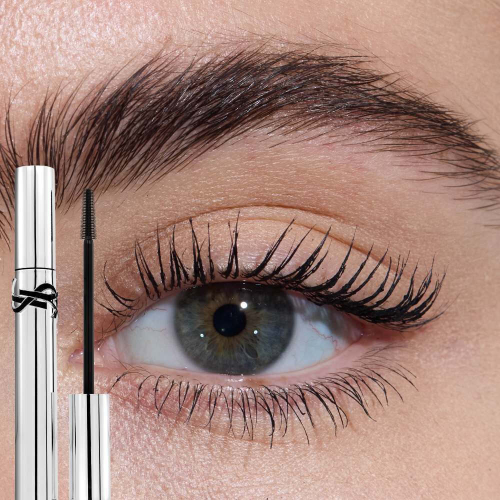 Close-up of an eye with light skin tone, showcasing lengthened lashes from YSL Lash Latex Mascara, with the silver tube and wand visible.
