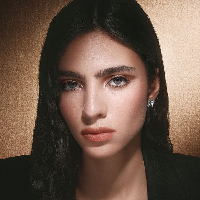 Close-up of model Loli Bahia with sparkling makeup and sleek black hair, set against a shimmering gold background.