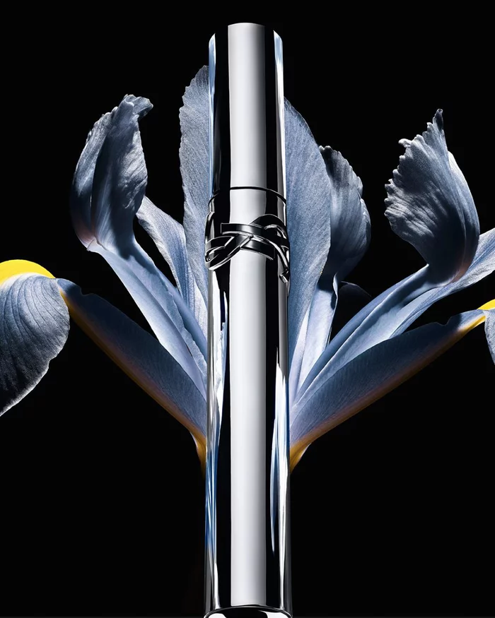 YSL Lash Latex Mascara, sleek silver tube with logo, elegantly placed within a blue iris flower on a dark background.