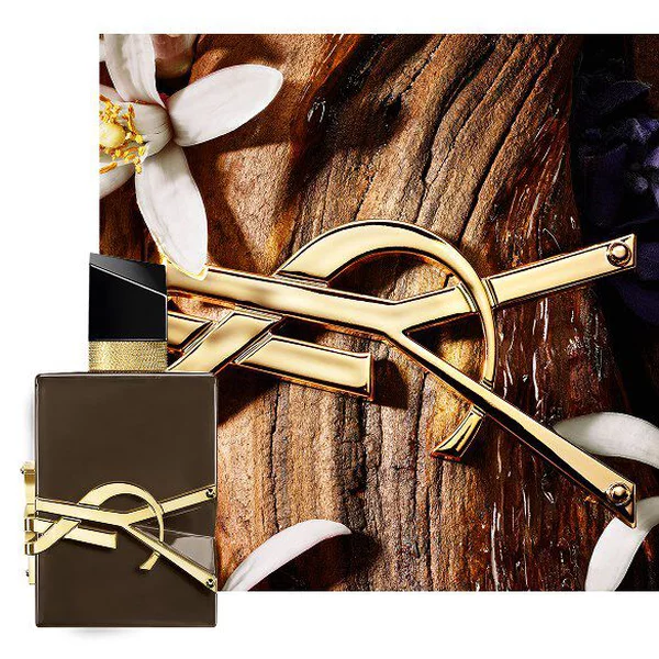 Libre Santal Couture glazed brown bottle on sandalwood bark with golden Cassandre and white flowers.