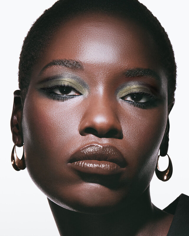 Awar, deep skin tone, with a smokey dawn eye look featuring green and gold, and glossy brown lips.