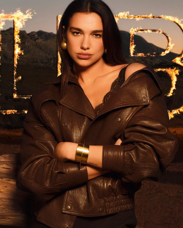 Dua Lipa in a brown leather jacket with large flaming LIBRE letters in a raw desert landscape.