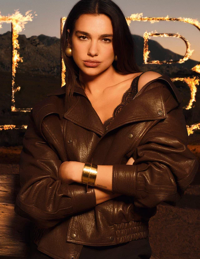 Dua Lipa in a brown leather jacket with large flaming LIBRE letters in a raw desert landscape.