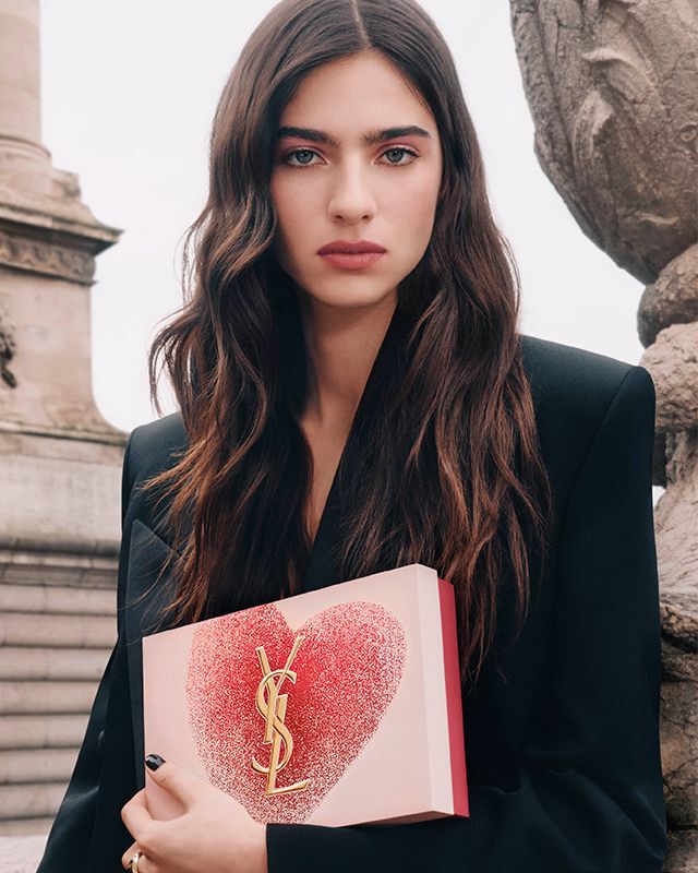YSL Beauty model Loli holding a pink heart-shaped gift set with a gold YSL logo, against a Parisian architectural background