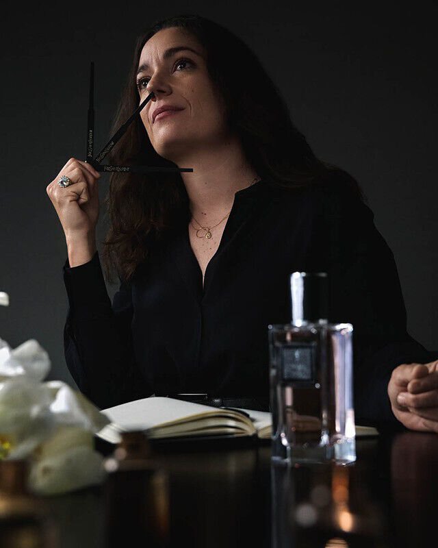 Perfumer Marie Salamagne, with dark hair, smelling YSL perfume blotters, with a fragrance bottle and white flowers nearby.