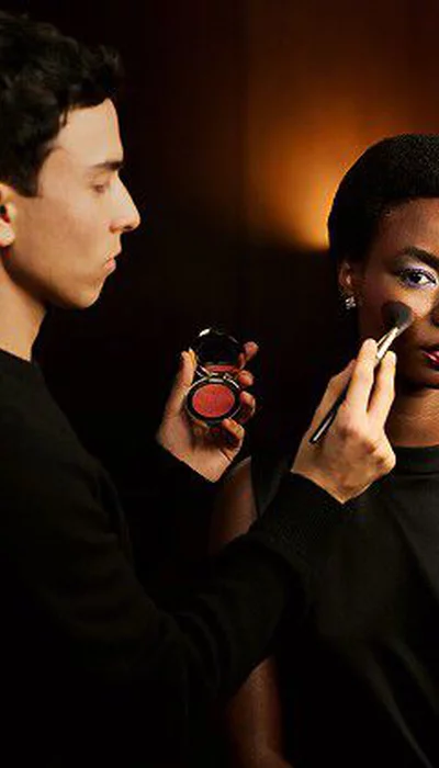 Makeup artist Sam Visser applies blush to a model with a bold eye look and glossy red lips, holding a YSL blush compact.