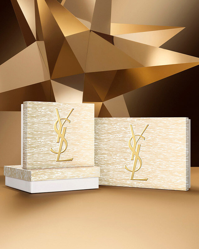 Holiday gift sets in gold tones with YSL logo, displayed on a geometric golden background.