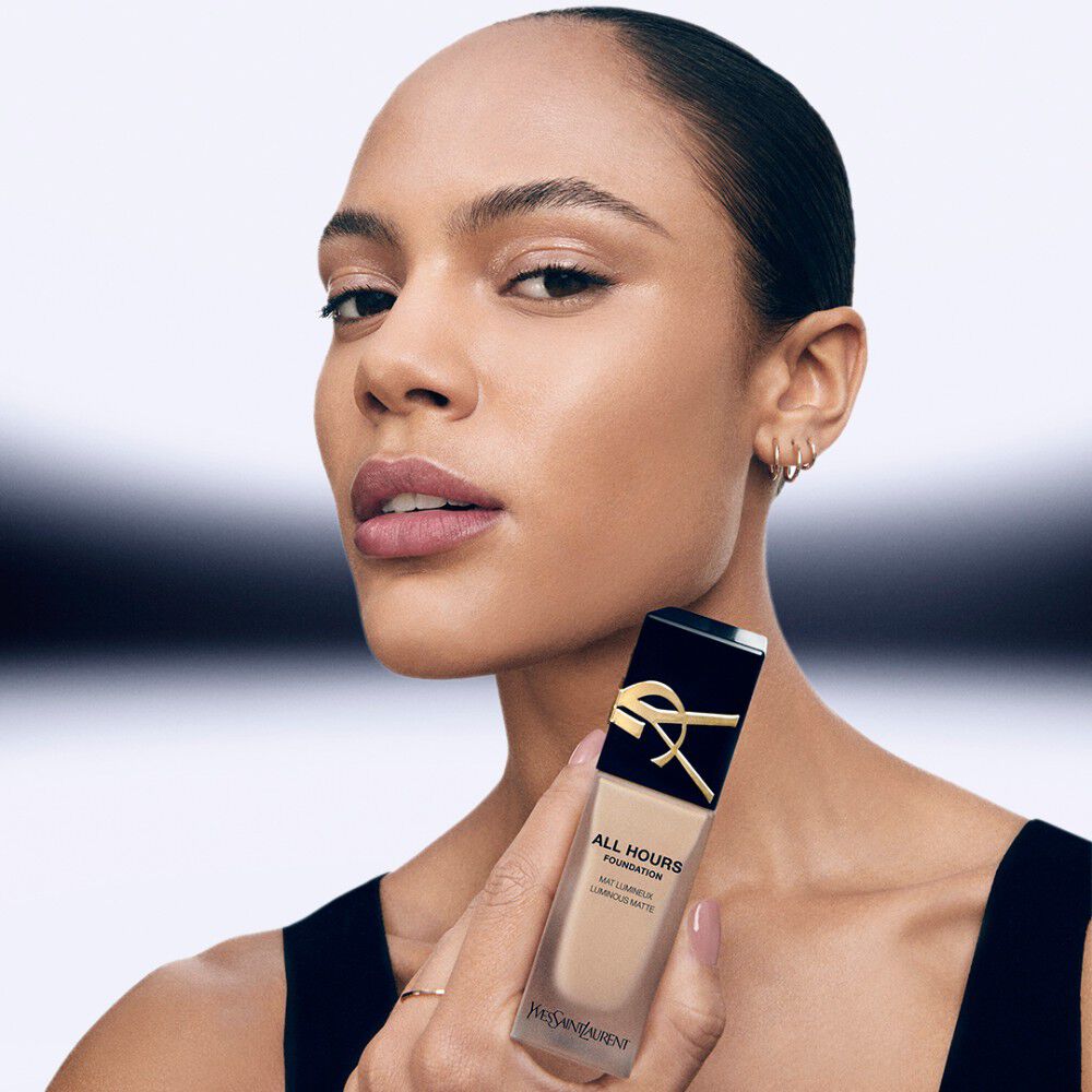 Model with medium skin tone holding YSL All Hours Foundation bottle, wearing a black top, close-up view.