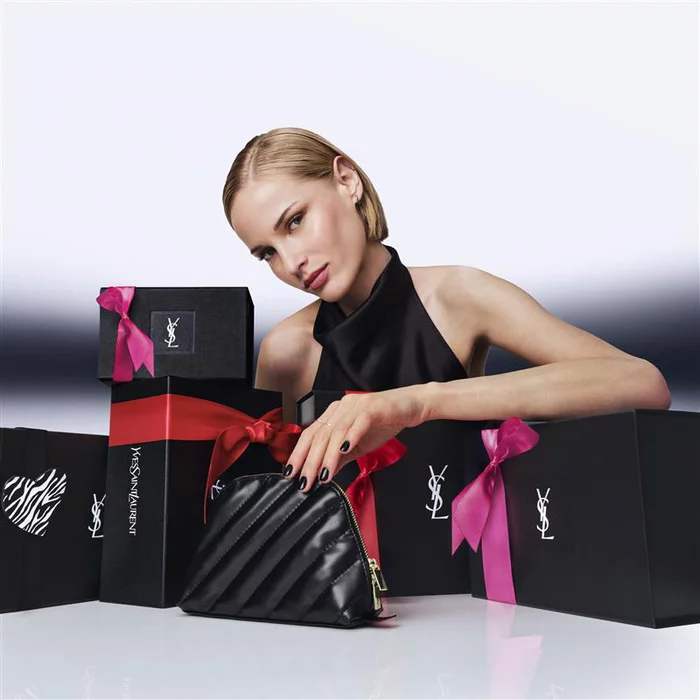 Arina, YSL Beauty model, surrounded by black YSL gift boxes with red and pink ribbons, holding a quilted YSL pouch.