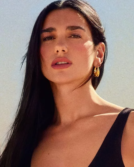 Dua Lipa, YSL Beauty ambassador, with flowing dark hair and a subtle tattoo, gazes directly at the camera in a desert setting.