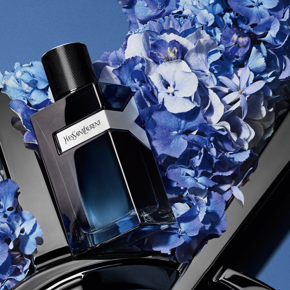 YSL Y Eau de Parfum bottle on a glossy black YSL logo, surrounded by vibrant blue hydrangeas, against a deep blue background.