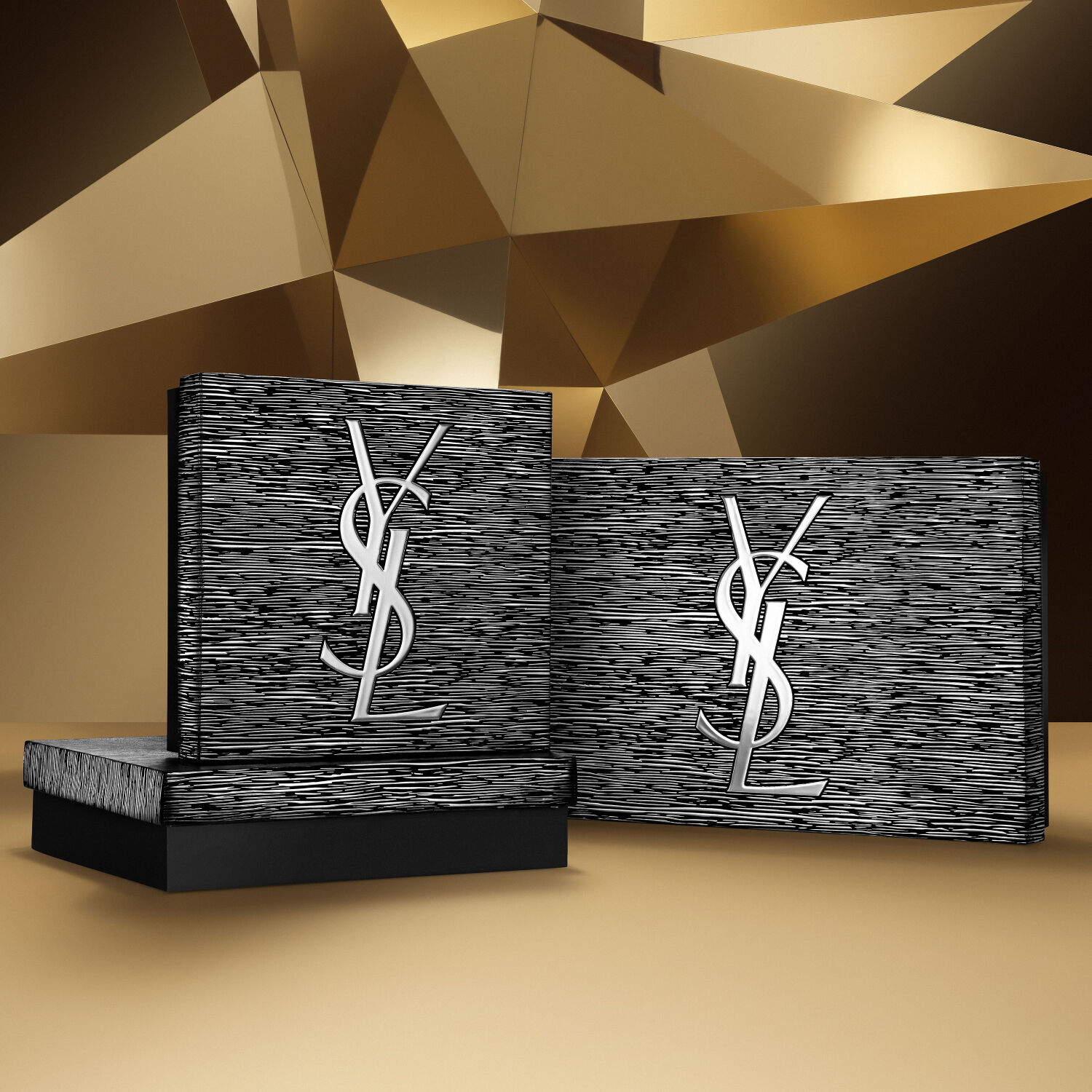 Two YSL Beauty Holiday gift sets in silver and black tones, featuring the YSL logo, on a golden geometric background.