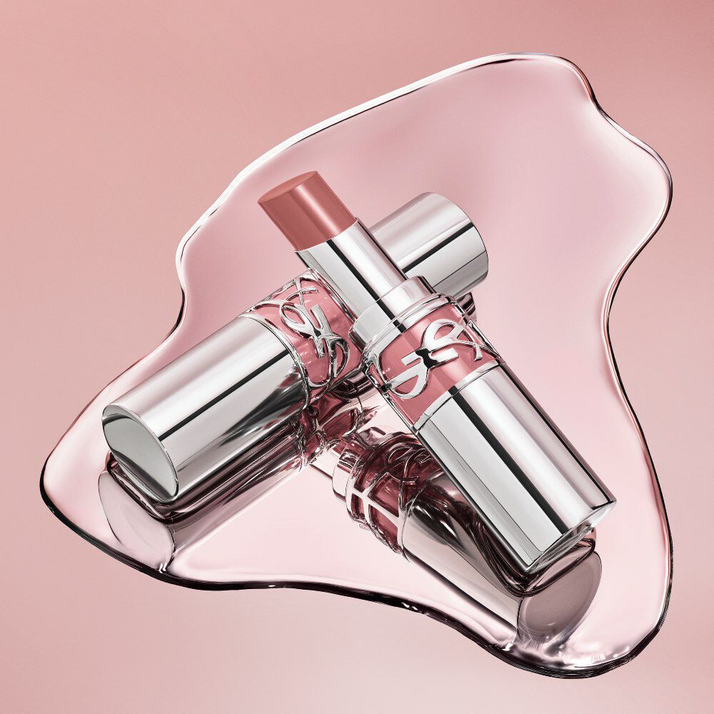Two silver YSL Loveshine lipstick tubes resting in a reflective, glossy puddle of pink liquid.