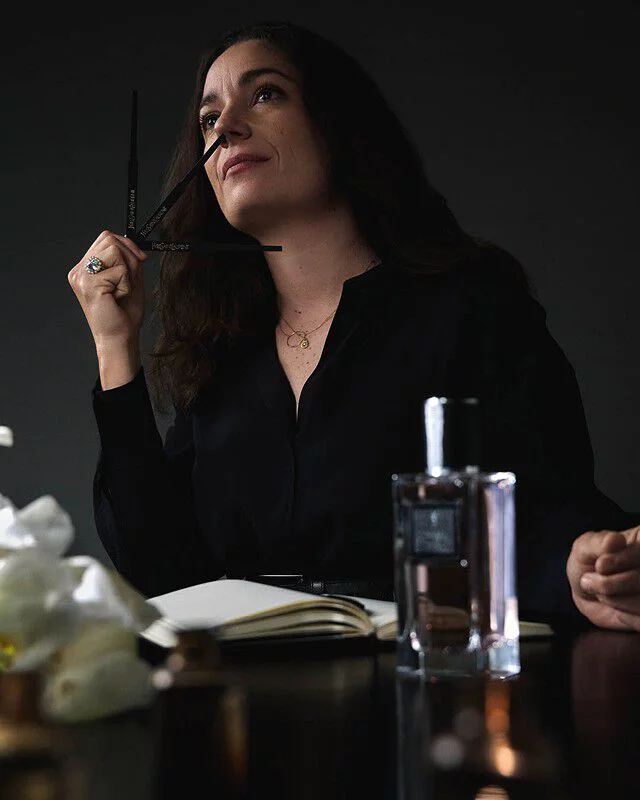Perfumer Marie Salamagne, with dark hair, smelling YSL perfume blotters, with a fragrance bottle and white flowers nearby.