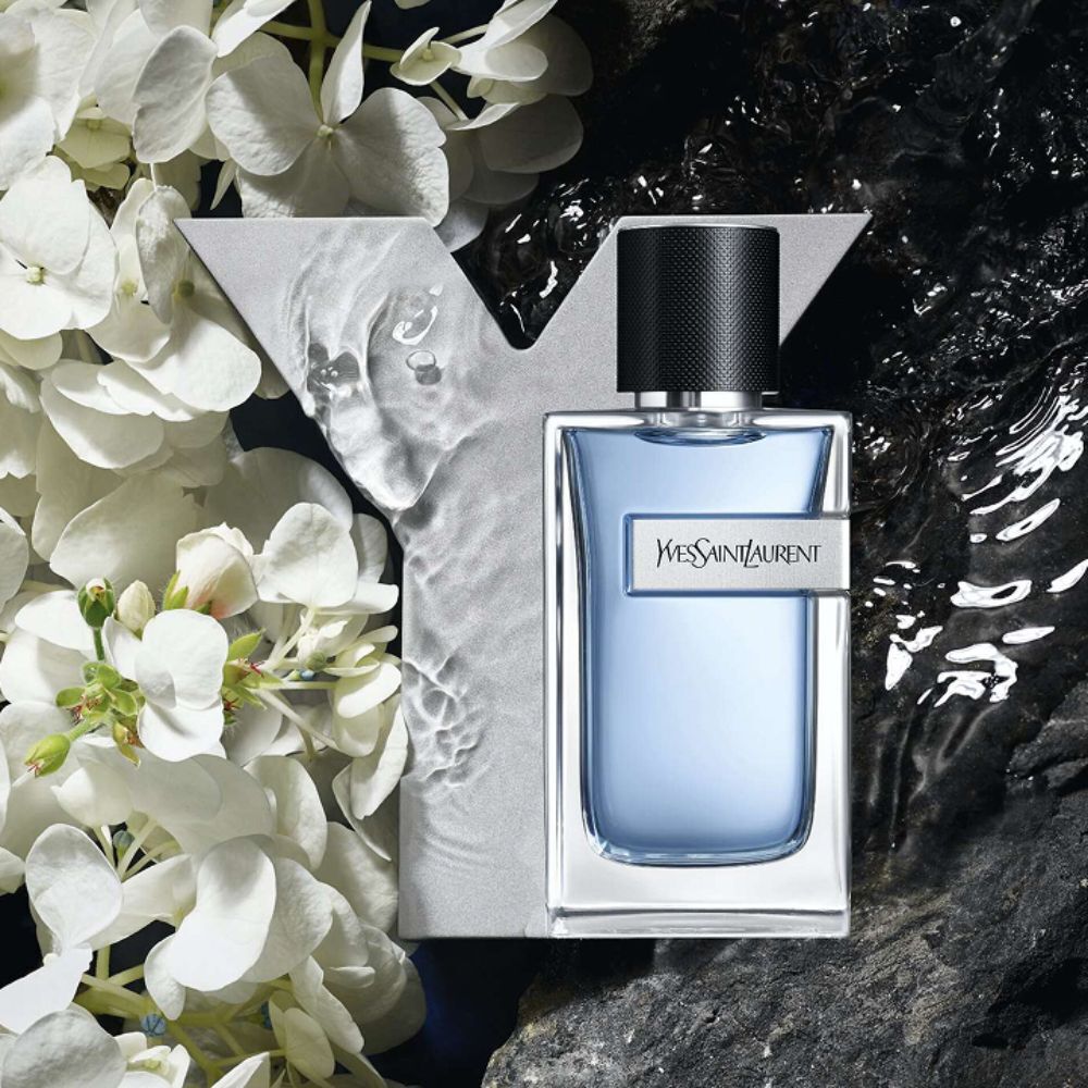 YSL Y Eau de Toilette bottle, light blue with silver Y logo, on dark rocks   with water splashes and elegant white flowers. 