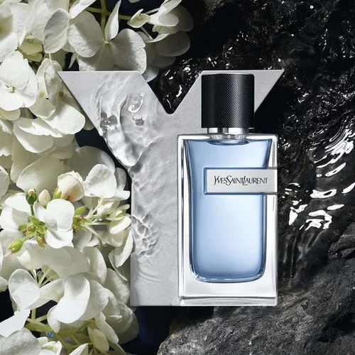 YSL Y Eau de Toilette bottle, light blue with silver Y logo, on dark rocks with water splashes and elegant white flowers.