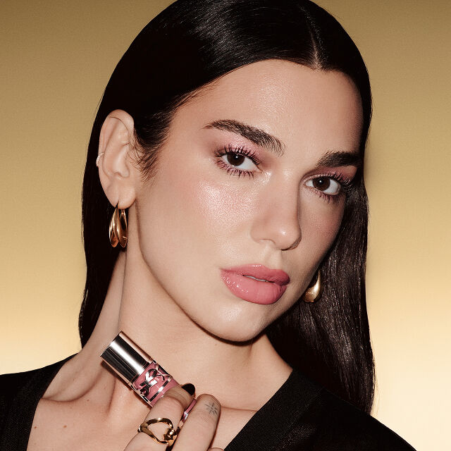 Dua Lipa, YSL Beauty ambassador, holding YSL Loveshine Plumping Lip Oil in a silver tube, on a gold gradient background.