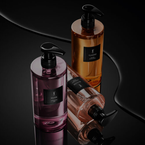 Le Vestiaire des Parfums Bath & Body Collection: three pump bottles—pink (Muse), amber (Tuxedo), and nude (Blouse)—on a glossy black surface with fluid reflections.