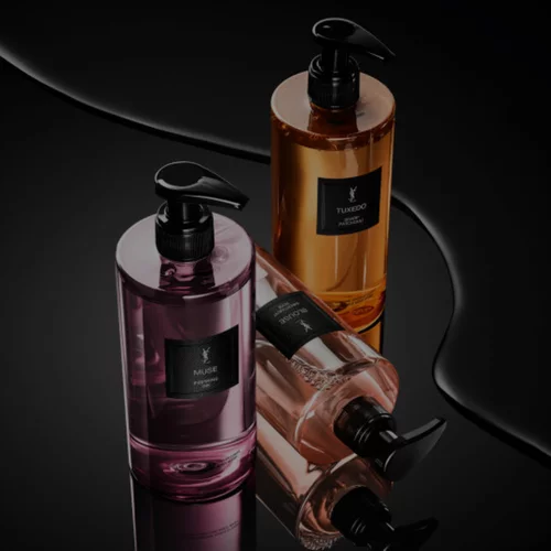 Le Vestiaire des Parfums Bath & Body Collection: three pump bottles—pink (Muse), amber (Tuxedo), and nude (Blouse)—on a glossy black surface with fluid reflections.