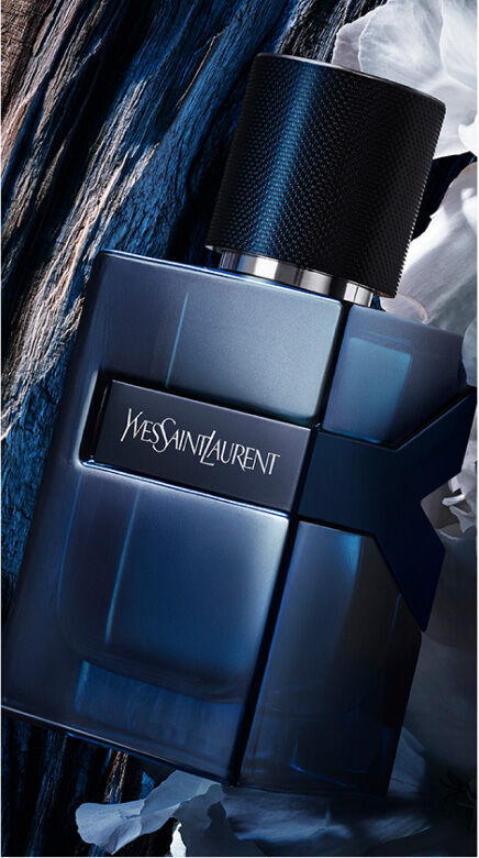 YSL Y L'Elixir bottle, dark blue with silver Y logo and textured cap, on a  dark wooden background with soft white petals.