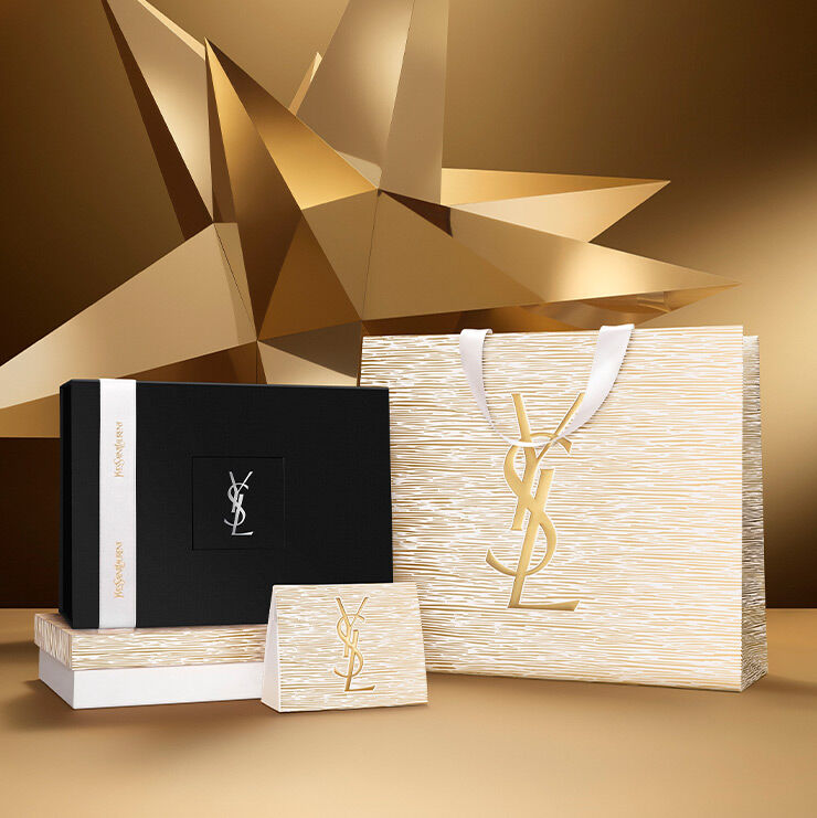Holiday gift set featuring a gold bag with YSL Beauty logo and a black box tied with a white ribbon, displayed on a starry gold-toned background.