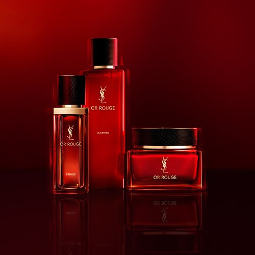 YSL Or Rouge L'Huile, La Lotion, and La Creme Essentielle, three red and gold skincare products, on a reflective red background.