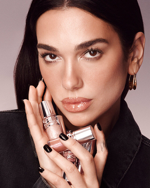 Dua Lipa, YSL Beauty ambassador, holding two YSL LOVESHINE Plumping Lip Oil Glosses, with a subtle gradient background.