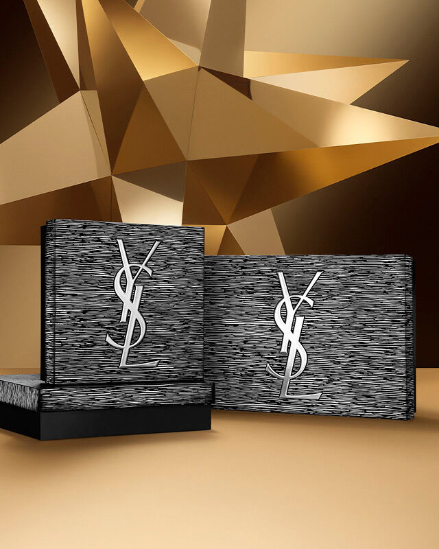 Two YSL Beauty Holiday gift sets in silver and black tones, featuring the YSL logo, on a golden geometric background.