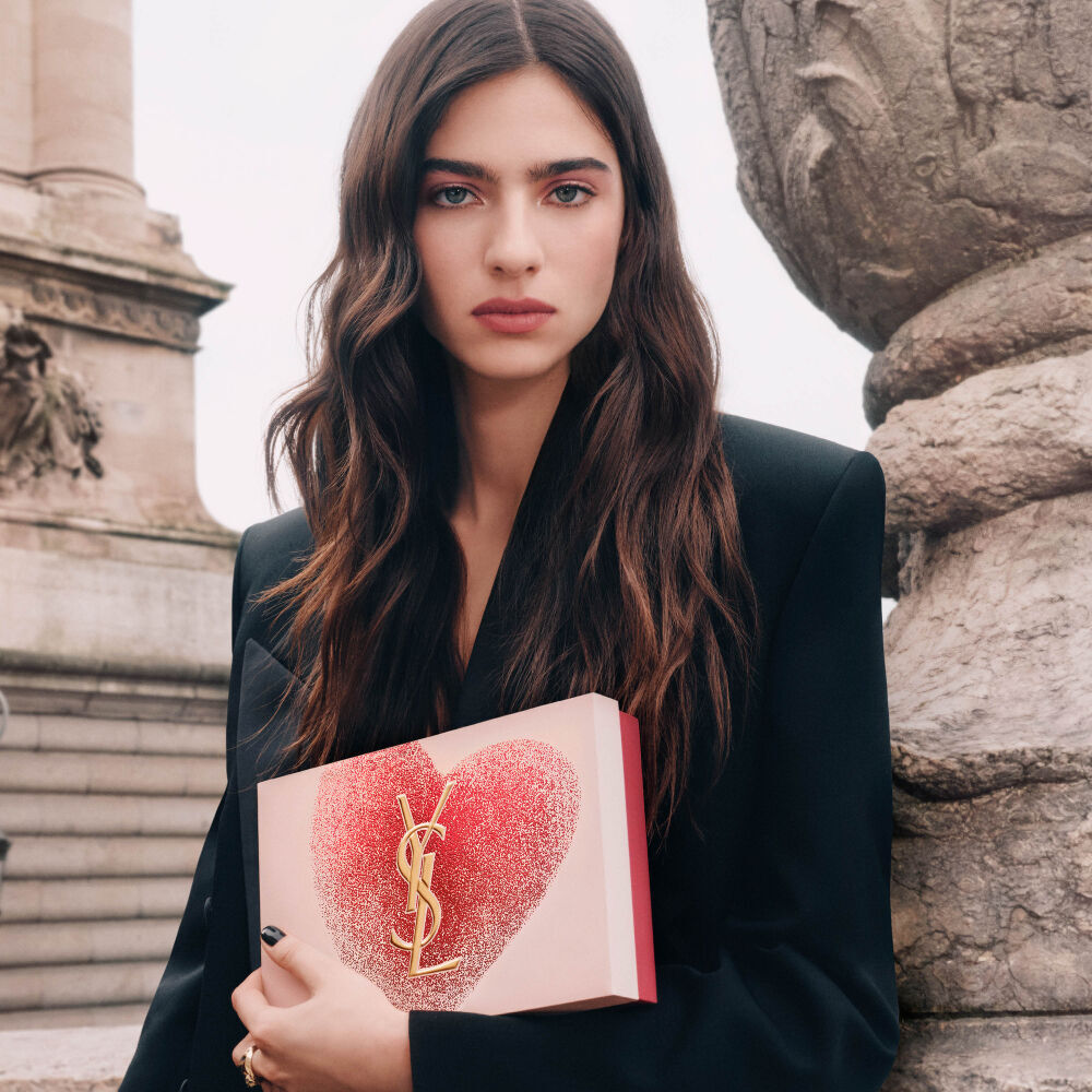 YSL Beauty model Loli holding a pink heart-shaped gift set with a gold YSL logo, against a Parisian architectural background