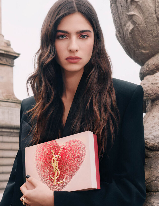 YSL Beauty model Loli holding a pink heart-shaped gift set with a gold YSL logo, against a Parisian architectural background