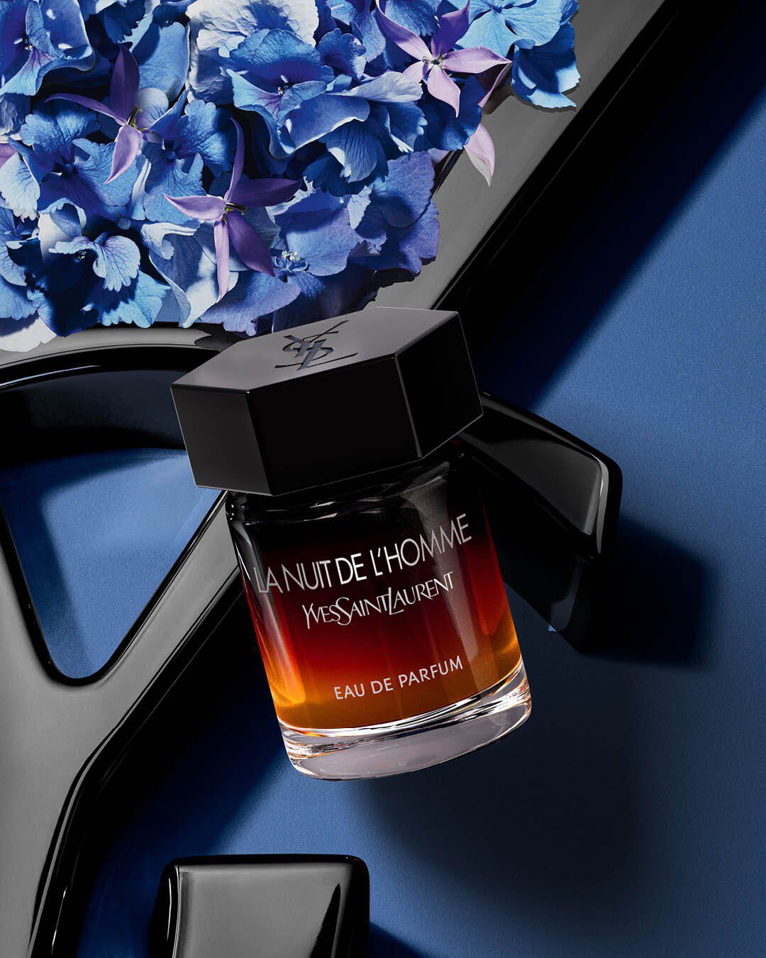 YSL La Nuit de l'Homme EDP bottle on a glossy black YSL logo, with vibrant blue hydrangeas, against a deep blue background.