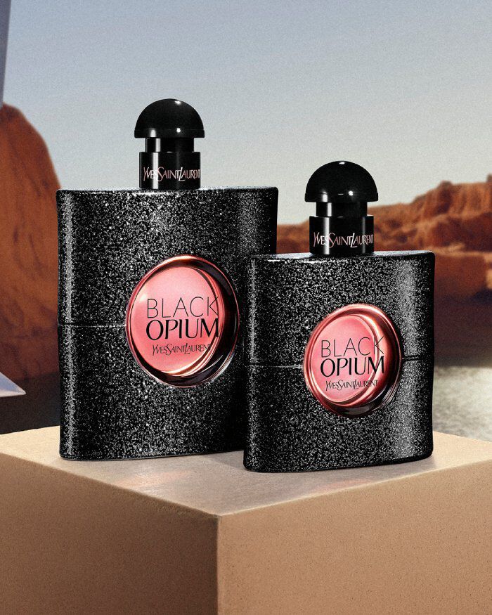 YSL Black Opium EDP 100ml and 50ml bottles, glittery black with pink labels, on a pedestal in a desert landscape.