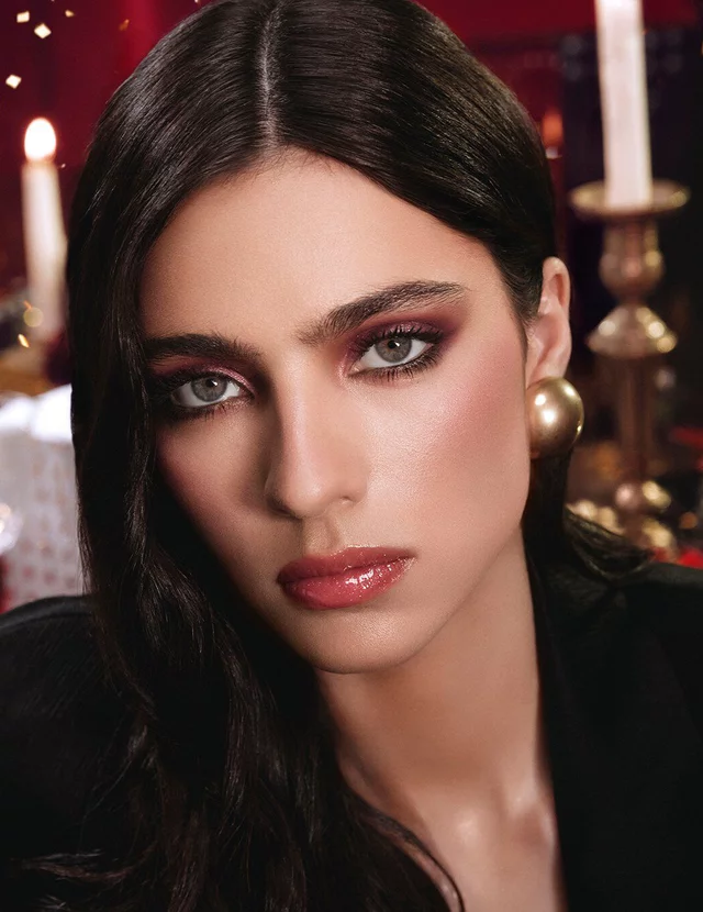 Model wearing a black suit and glam look, seated in a festive candlelit setting with YSL gift boxes.