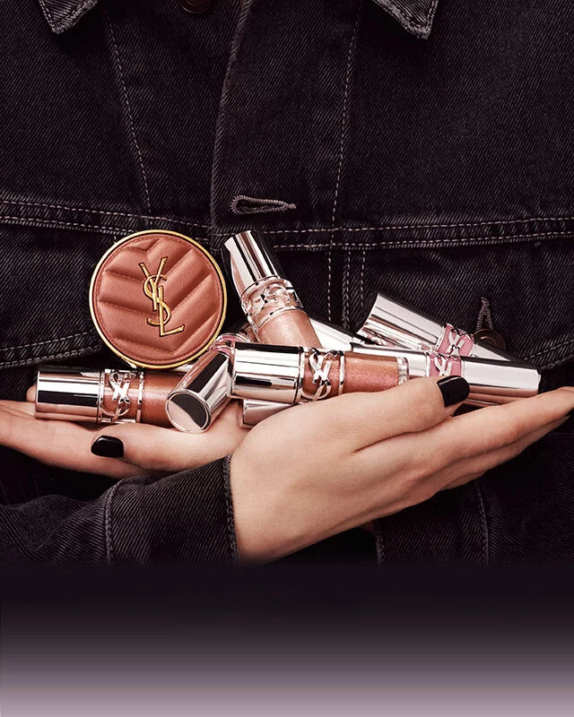 Hands holding YSL LOVESHINE Plumping Lip Oil Gloss and Make Me Blush, showcasing luxury beauty products.
