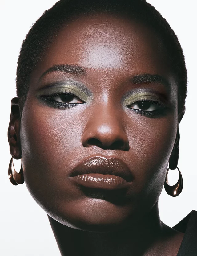 Awar, deep skin tone, with a smokey dawn eye look featuring green and gold, and glossy brown lips.