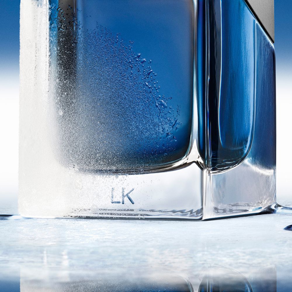 Close-up of the frosted base of a YSL Y Eau de Toilette Intense Iced  Cologne bottle, with "LK" engraving, on a cool blue surface.