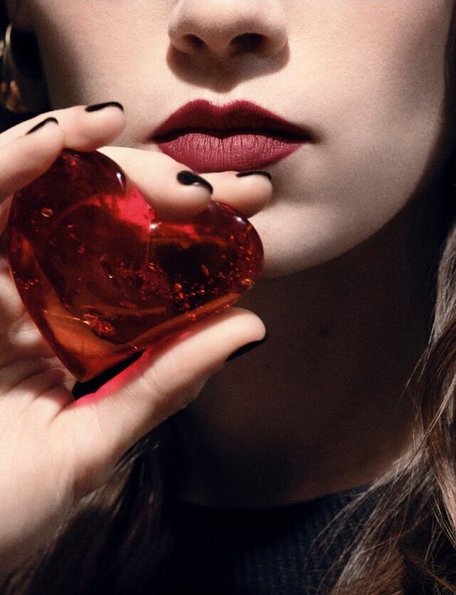 Close-up of a model's lips and hand holding a translucent ruby-red heart jewel for YSL COEUR Eau de Parfum.