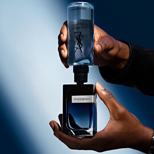 Hands refilling a YSL Y fragrance bottle with "THE REFILL LA RECHARGE" bottle, highlighting sustainable luxury.