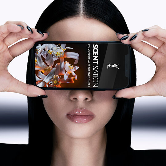 Model with sleek black nails holding a smartphone showcasing the YSL fragrance app, close-up view.