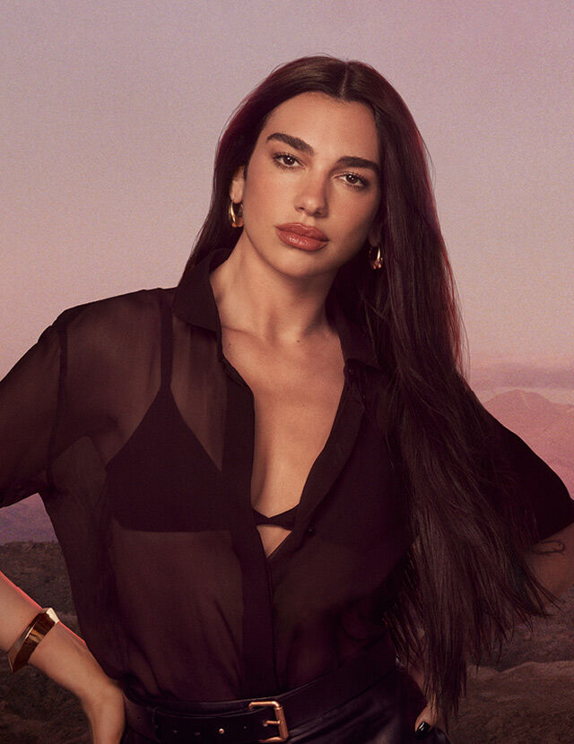 Dua Lipa with long dark hair wearing a black blouse, standing against a pink sunset landscape background.