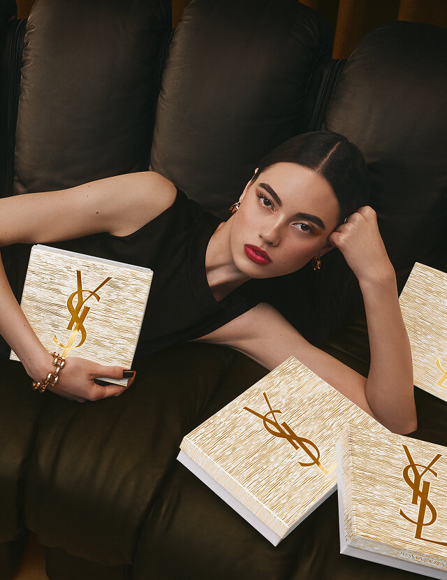Model reclining on a black sofa surrounded by YSL Holiday gift sets in gold and white with YSL logo.