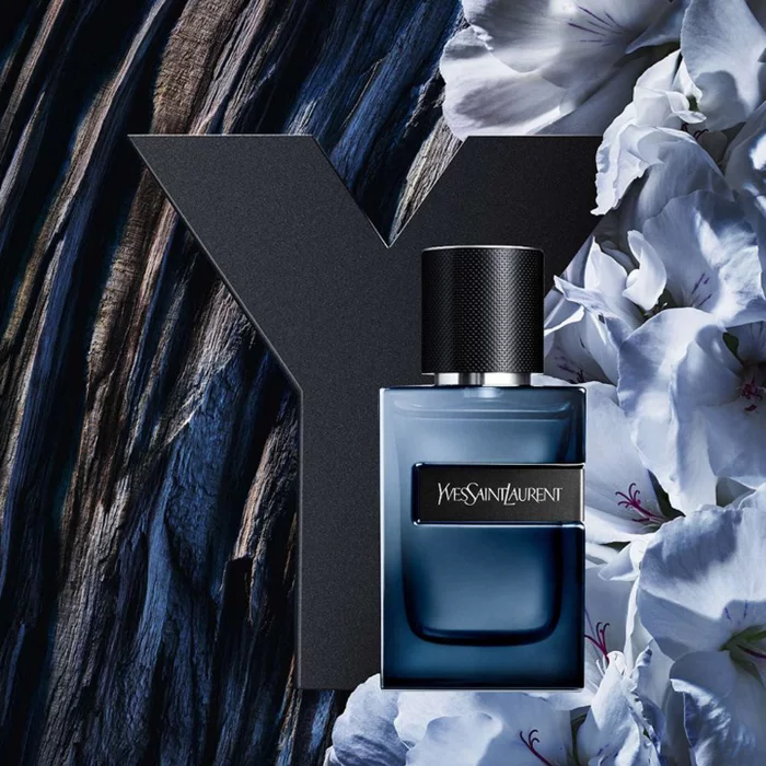 YSL Y L'Elixir bottle, dark blue with black Y logo and textured cap, on a dark wooden background with elegant white flowers.