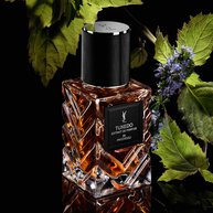 YSL LVDP Tuxedo Extrait de Parfum bottle, framed by lush green patchouli leaves and delicate purple flowers on black.