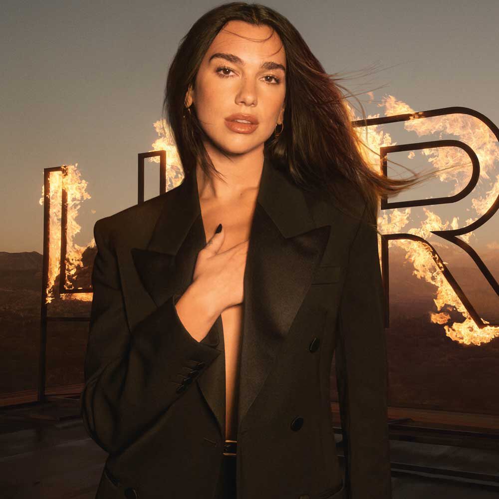 Dua Lipa in a black blazer with flames in the background.