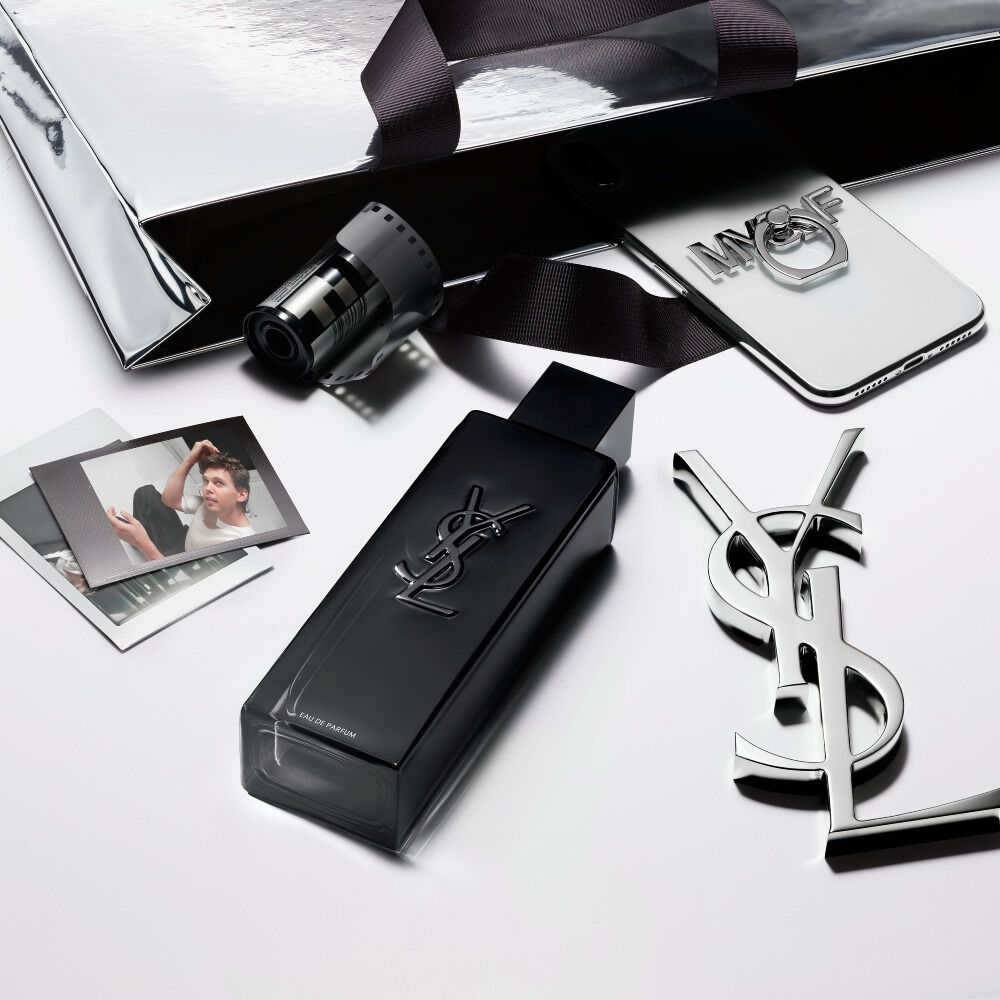 A top-down view of the MYSLF bottle, a black YSL gift bag, polaroid photos, and a silver YSL logo charm arranged on a white surface.
