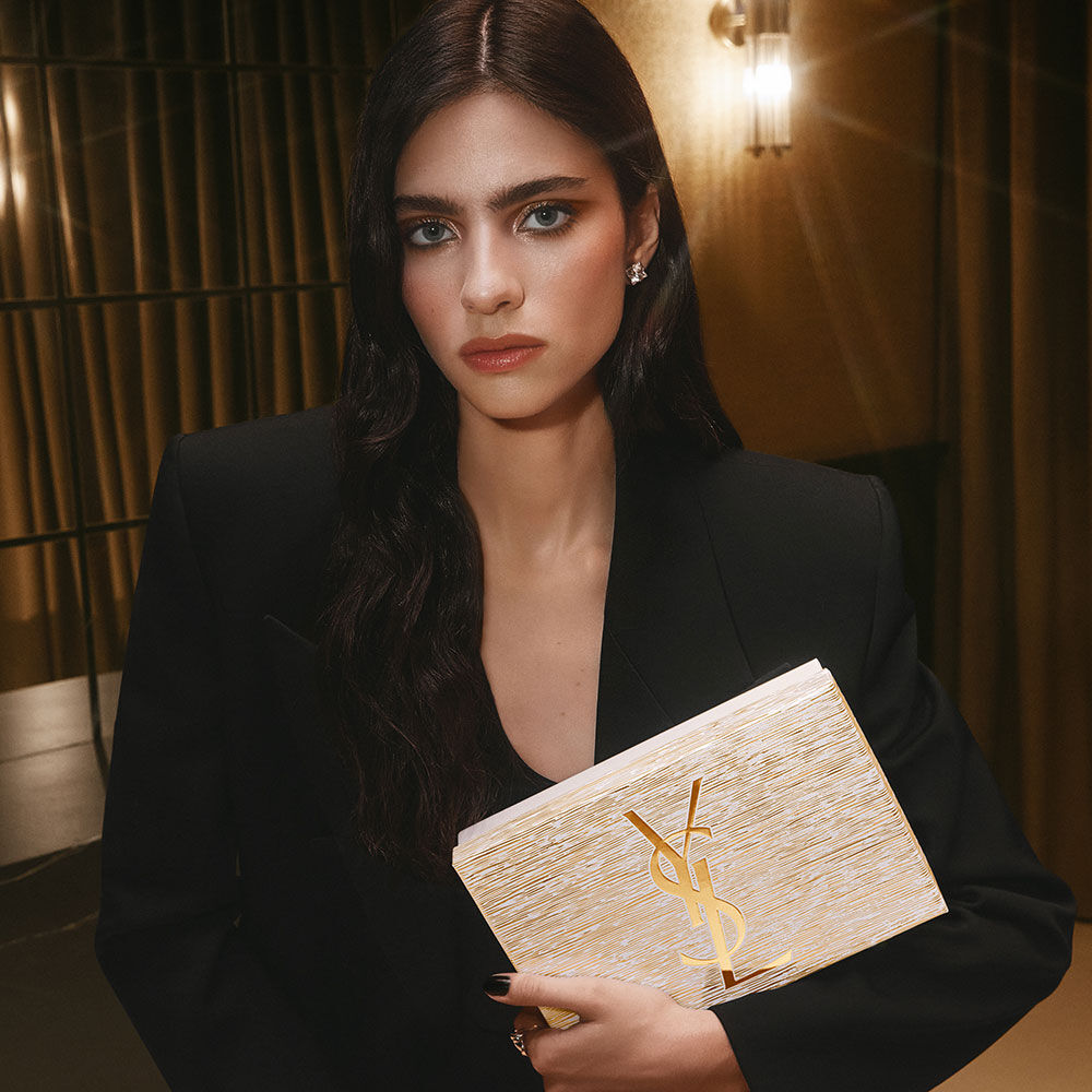 Model Loli Bahia holding a golden YSL Holiday gift set, wearing a black blazer and sophisticated makeup.