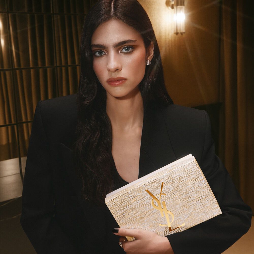 Model Loli Bahia holding a golden YSL Holiday gift set, wearing a black blazer and sophisticated makeup.