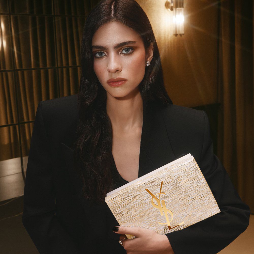 Model Loli Bahia holding a golden YSL Holiday gift set, wearing a black blazer and sophisticated makeup.