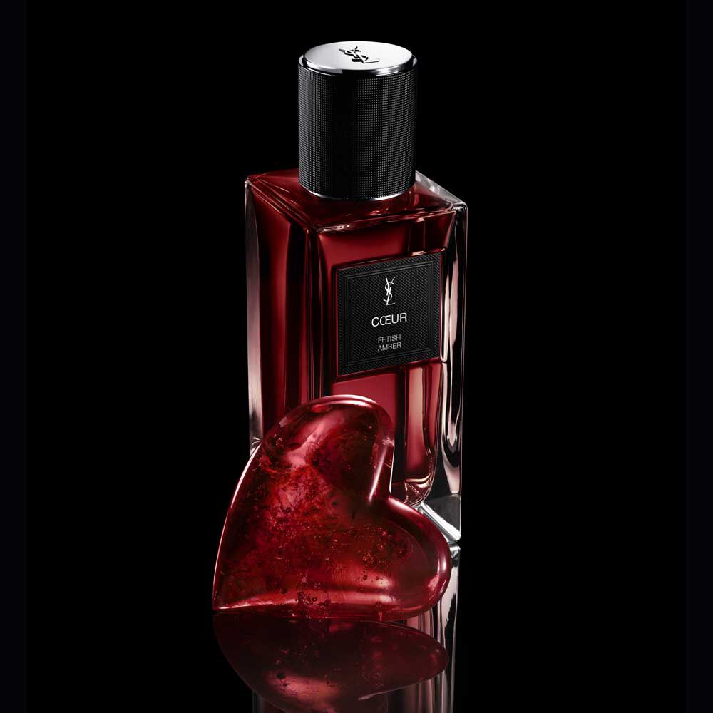 COEUR Eau de Parfum by YSL bottle next to a ruby-red heart jewel on a reflective black surface.