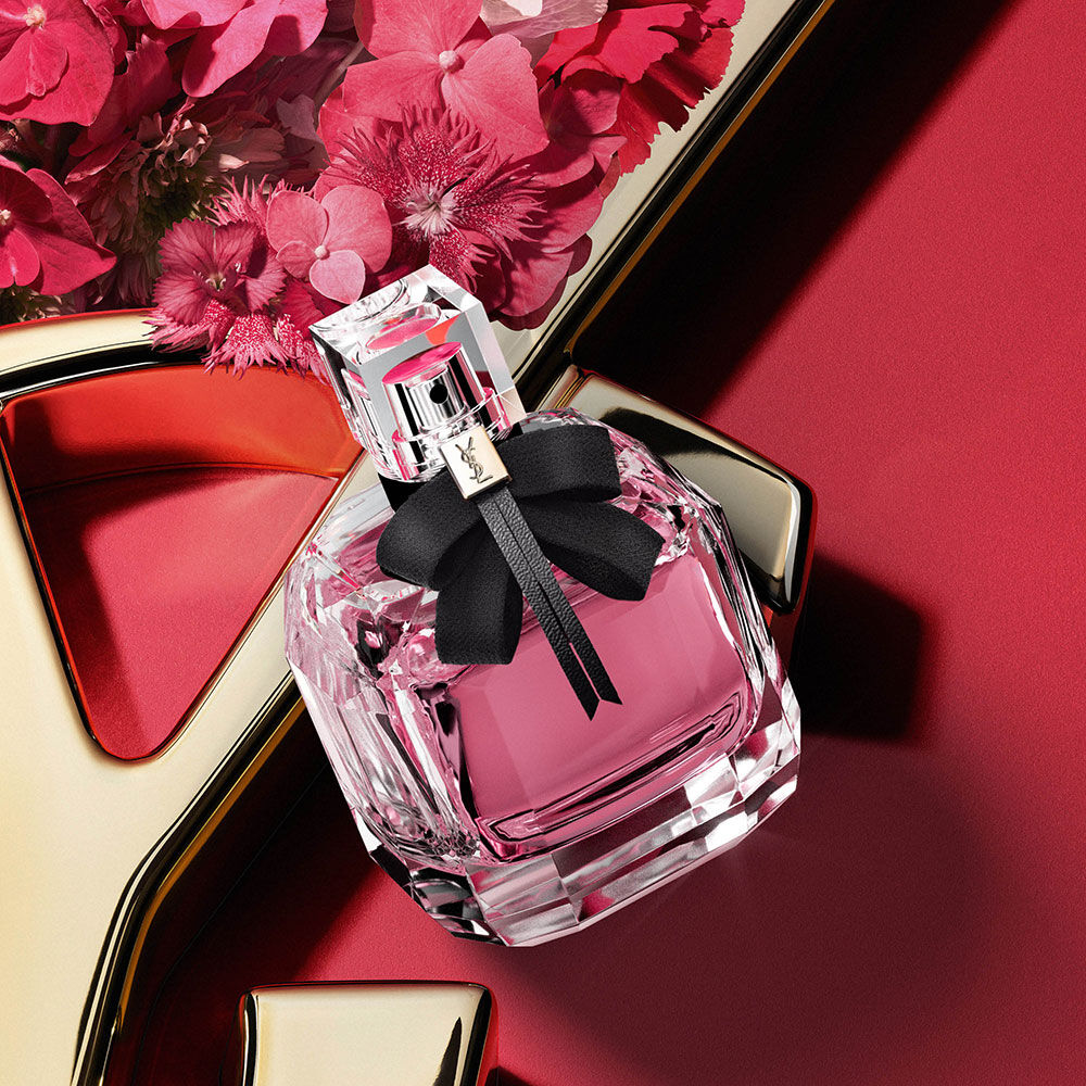 YSL Mon Paris Eau de Parfum bottle with pink fragrance and a black bow, styled with pink flowers on a red background with a gold YSL logo detail.